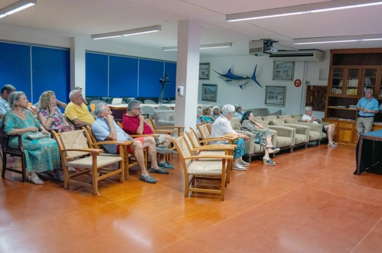 Grupo de personas mayores sentadas en una sala, escuchando a un hombre que está de pie en la esquina derecha, probablemente en una presentación o charla.