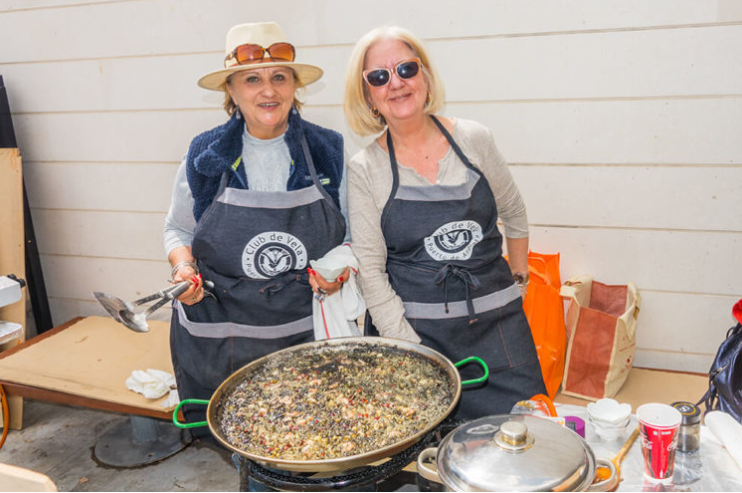 Dos mujeres mayores con gafas y aprons de un club de vela, preparando comida en una parrilla grande con ingredientes y utensilios, en un ambiente exterior de madera.