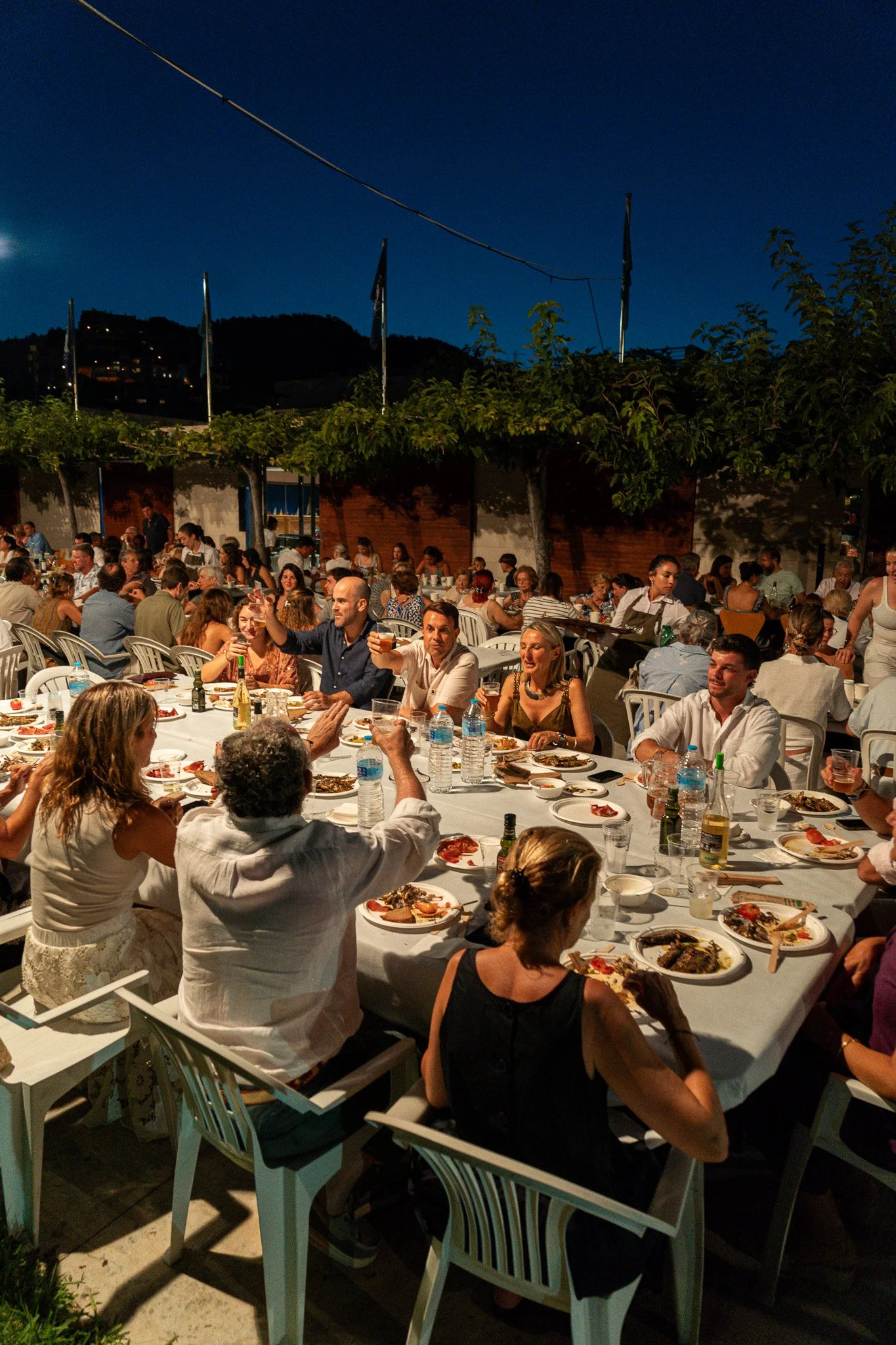 Una reunión social al aire libre por la noche con muchas personas sentadas en mesas largas con comida y bebidas, en un lugar rodeado de árboles y con un cielo oscuro.