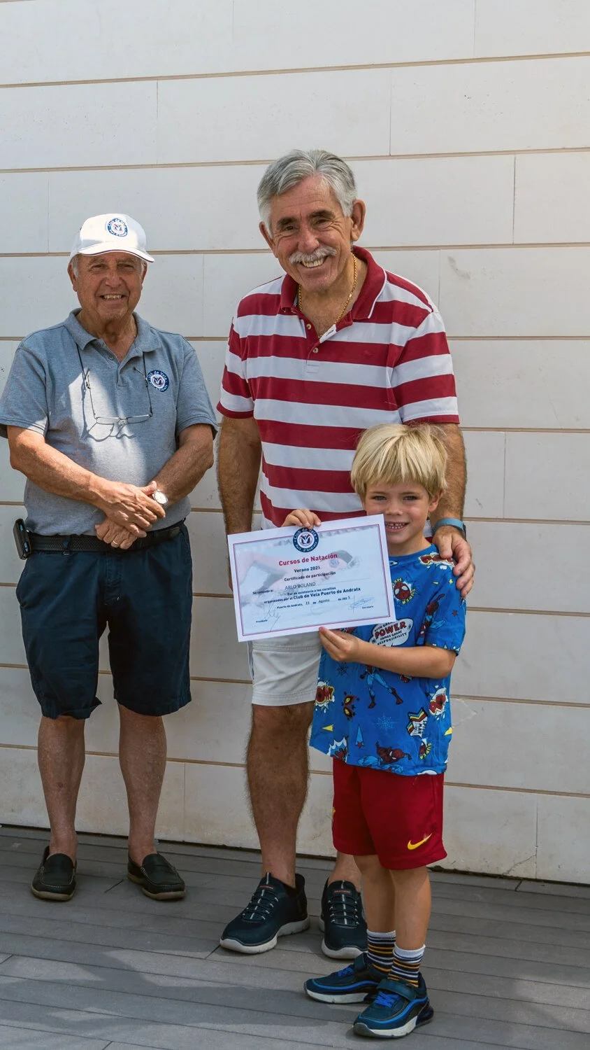 Un niño con pantalones cortos rojos y camiseta azul recibe un certificado de natación de manos de un hombre con camiseta de rayas rojas y blancas, mientras otro hombre en fondo vista con gorra observa. Todos sonríen y parecen en un evento de clausura