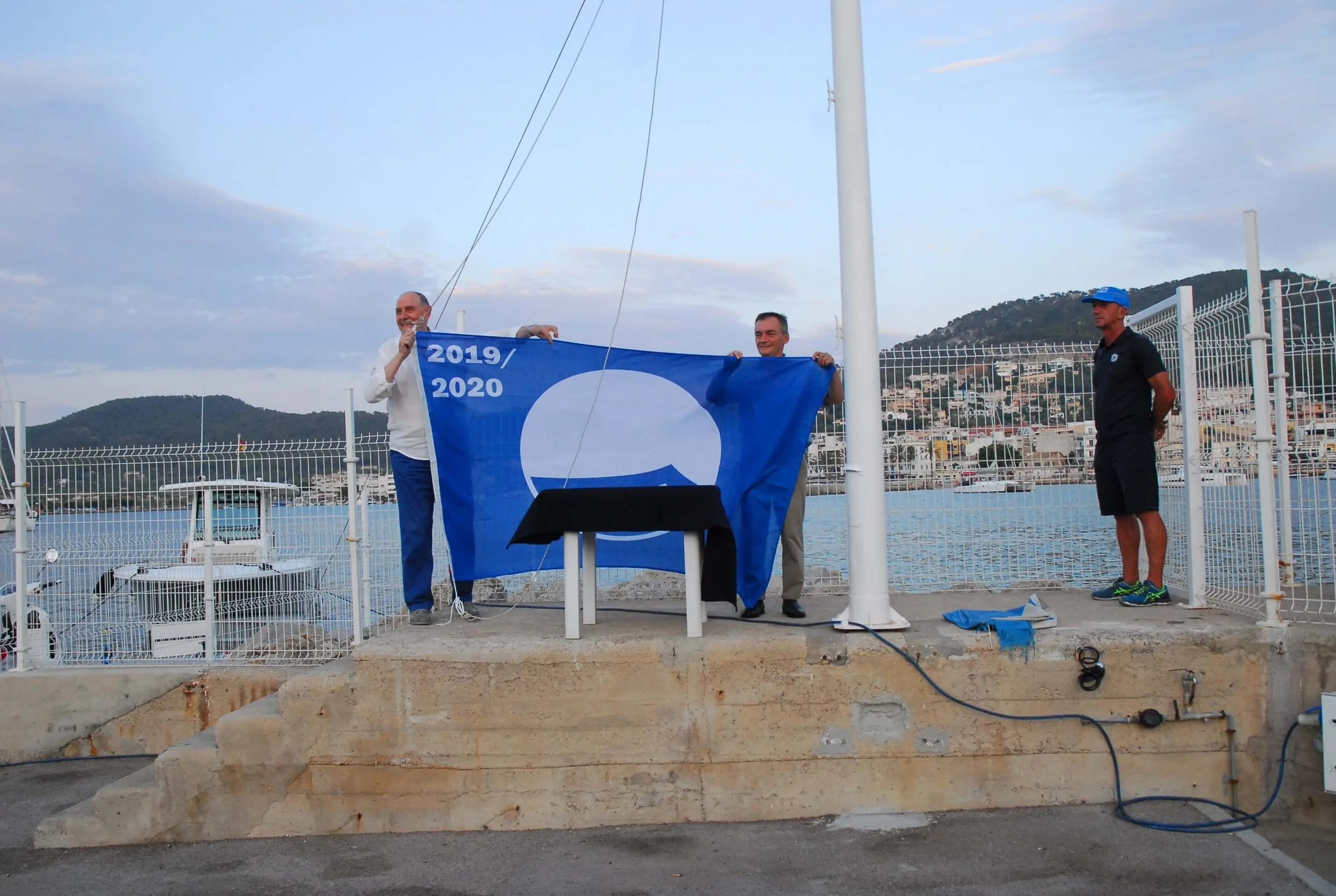 Tres hombres en una área abierta con vista a la ciudad y montañas, colocando una bandera azul con círculos blancos y los años 2019 y 2020, cerca de una cerca y algunos cables en el suelo.