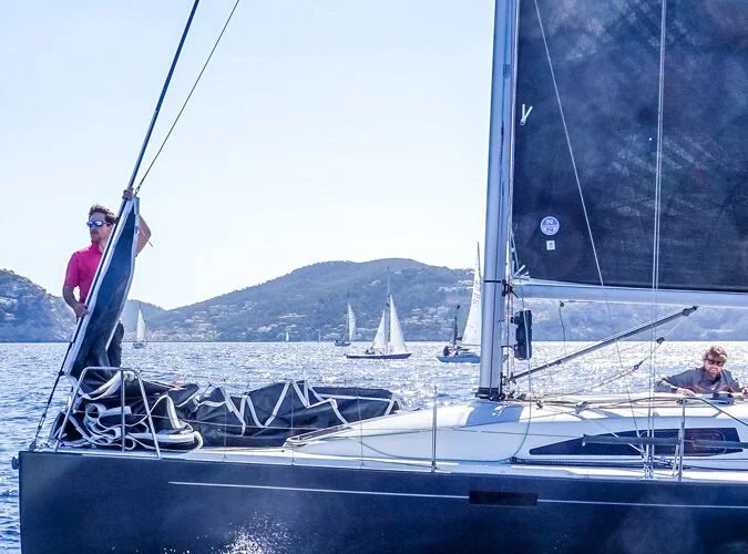 Dos personas en un velero negro navegando en el agua con varias embarcaciones en el fondo y una colina.