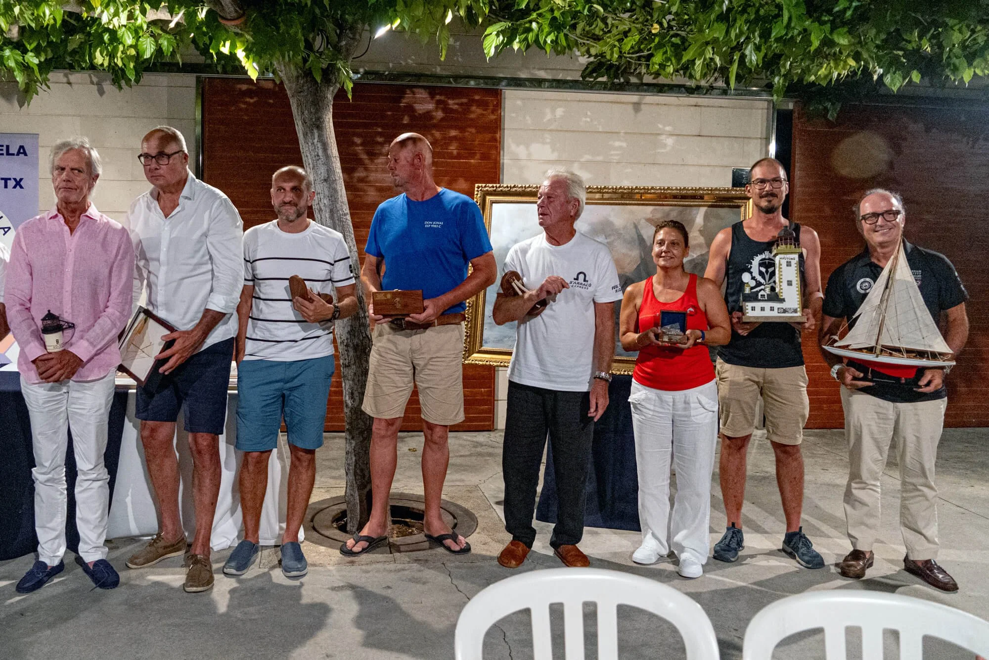 Grupo de nueve personas de pie frente a una pared, algunos sosteniendo objetos, en un evento social nocturno al aire libre con un árbol y un marco dorado en el fondo.