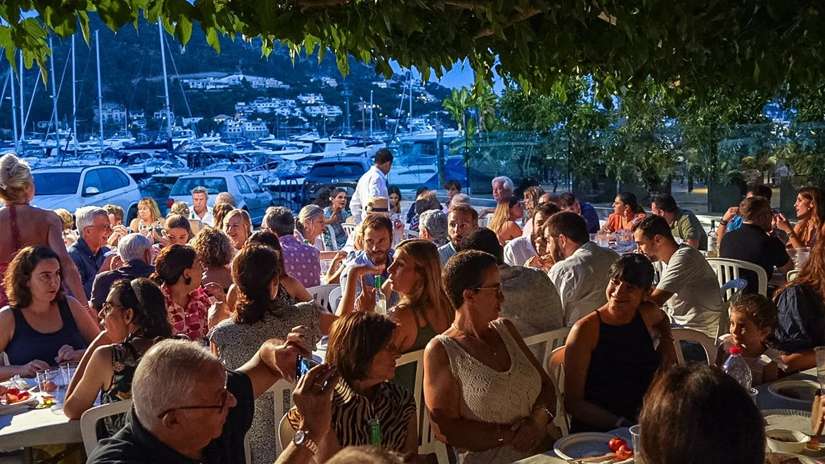 Personas en una comida al aire libre junto a un puerto con yates y montañas en el fondo, bajo sombra de árboles.