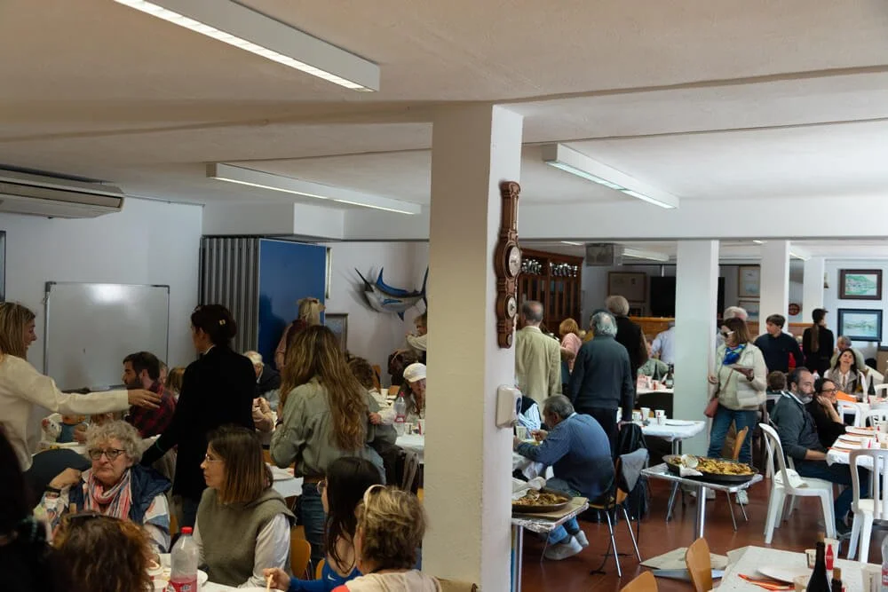 Una sala de restaurante concurrida con muchas personas sentadas en mesas, sirviendo comida y conversando, con decoraciones de temática marítima en la pared.