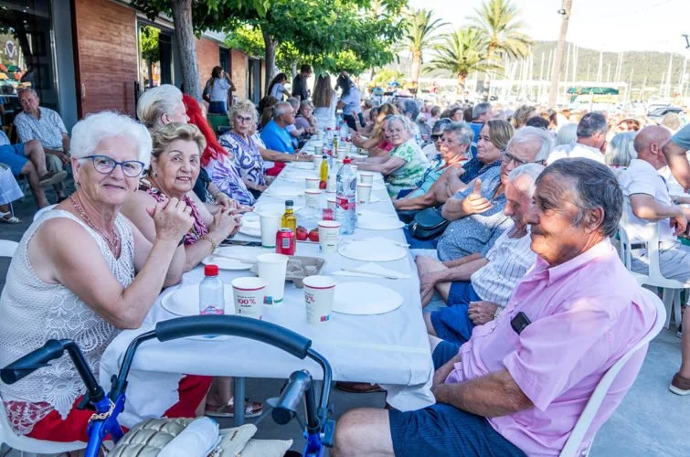 Grupo de personas mayores sentadas en una mesa larga al aire libre, participando en una comida o reunión social, con muchas palmeras y árboles al fondo.