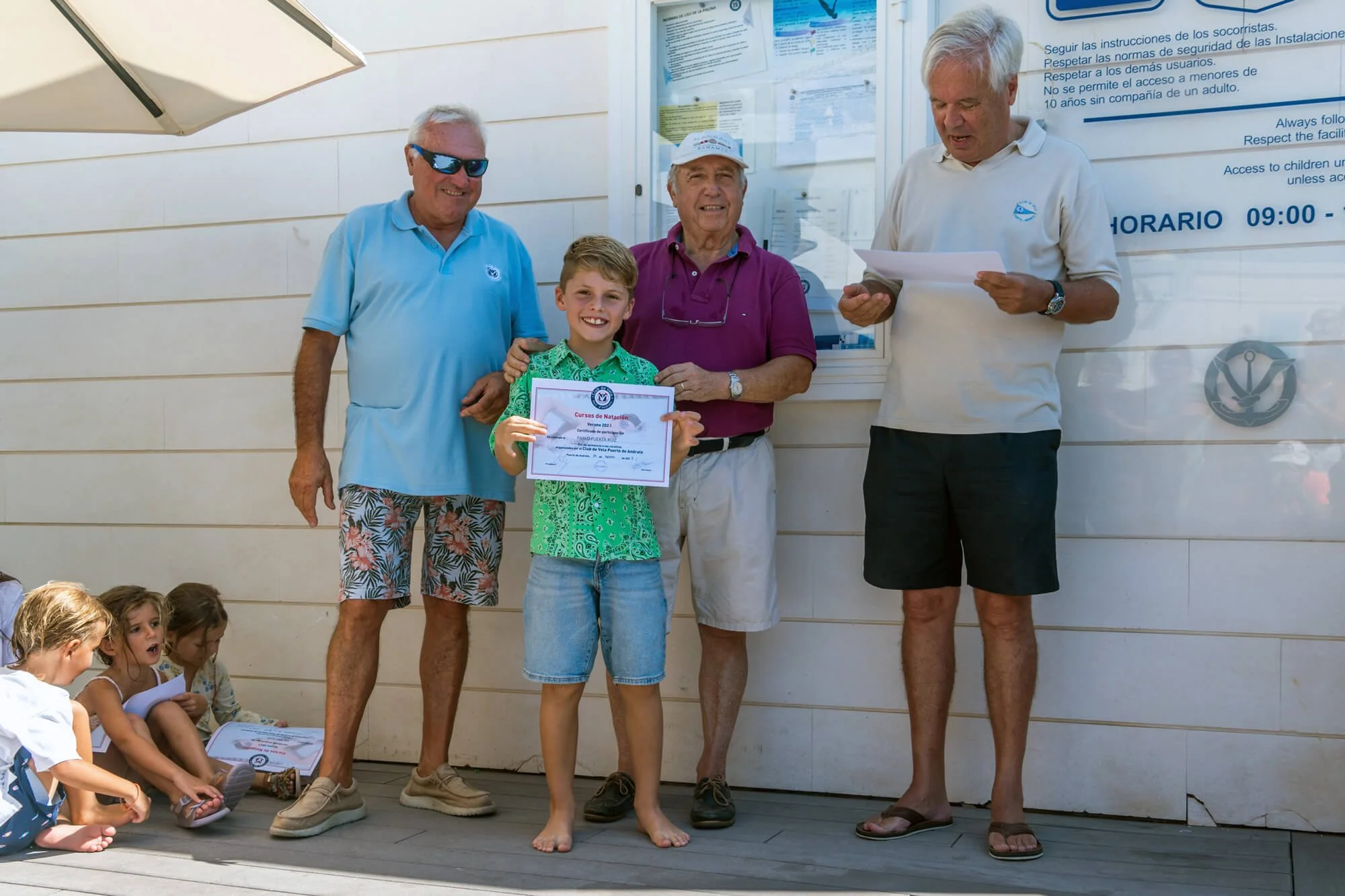 Niño sonriente recibe un certificado de natación en una ceremonia al aire libre, acompañado por tres adultos, mientras otros niños observan sentados a un lado.
