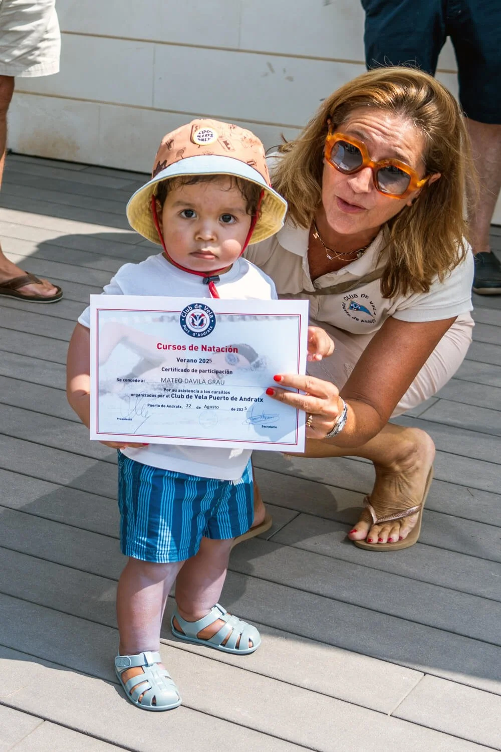 Niño pequeño con sombrero y camiseta blanca sostiene un certificado de participación en cursos de natación, acompañado por una mujer con gafas de sol anaranjadas, en una ceremonia al aire libre.
