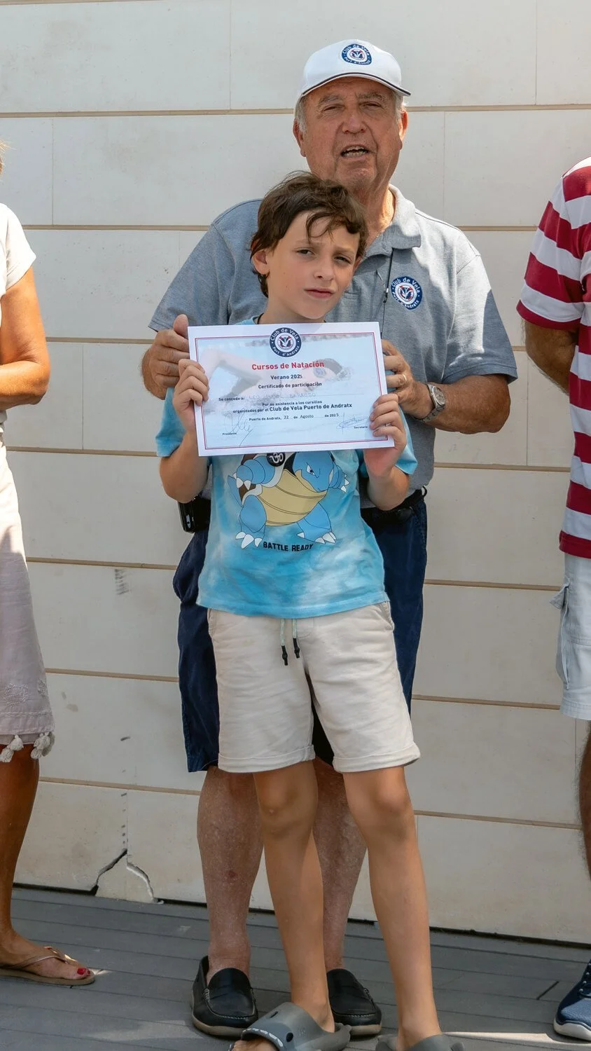 Niño sosteniendo un certificado de participación en un curso de natación, acompañado por un adulto en un evento al aire libre, en un muro de fondo.