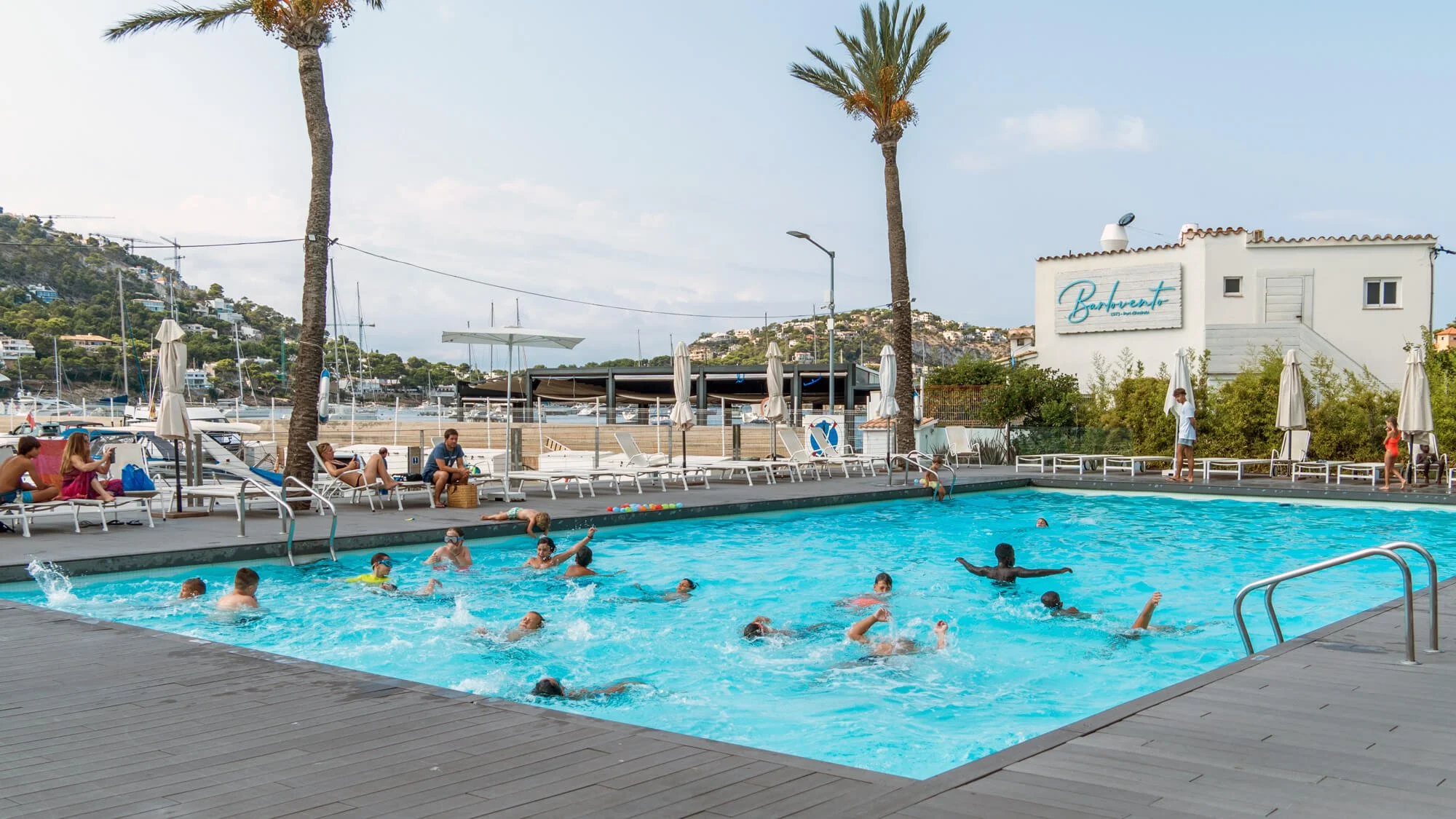 Personas disfrutando en una piscina con vista al mar en un día soleado, rodeada de sillones y sombrillas, con edificios y palmeras en el fondo.