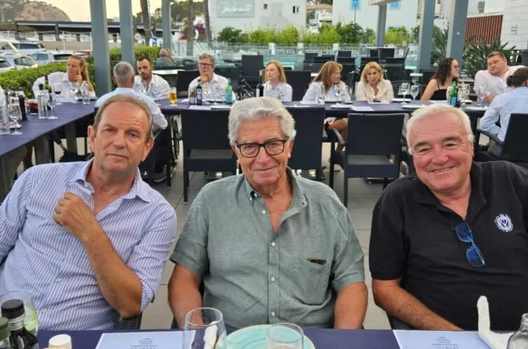 Tres hombres sentados en una mesa en un restaurante al aire libre, con varias personas en el fondo en una reunión o evento social.
