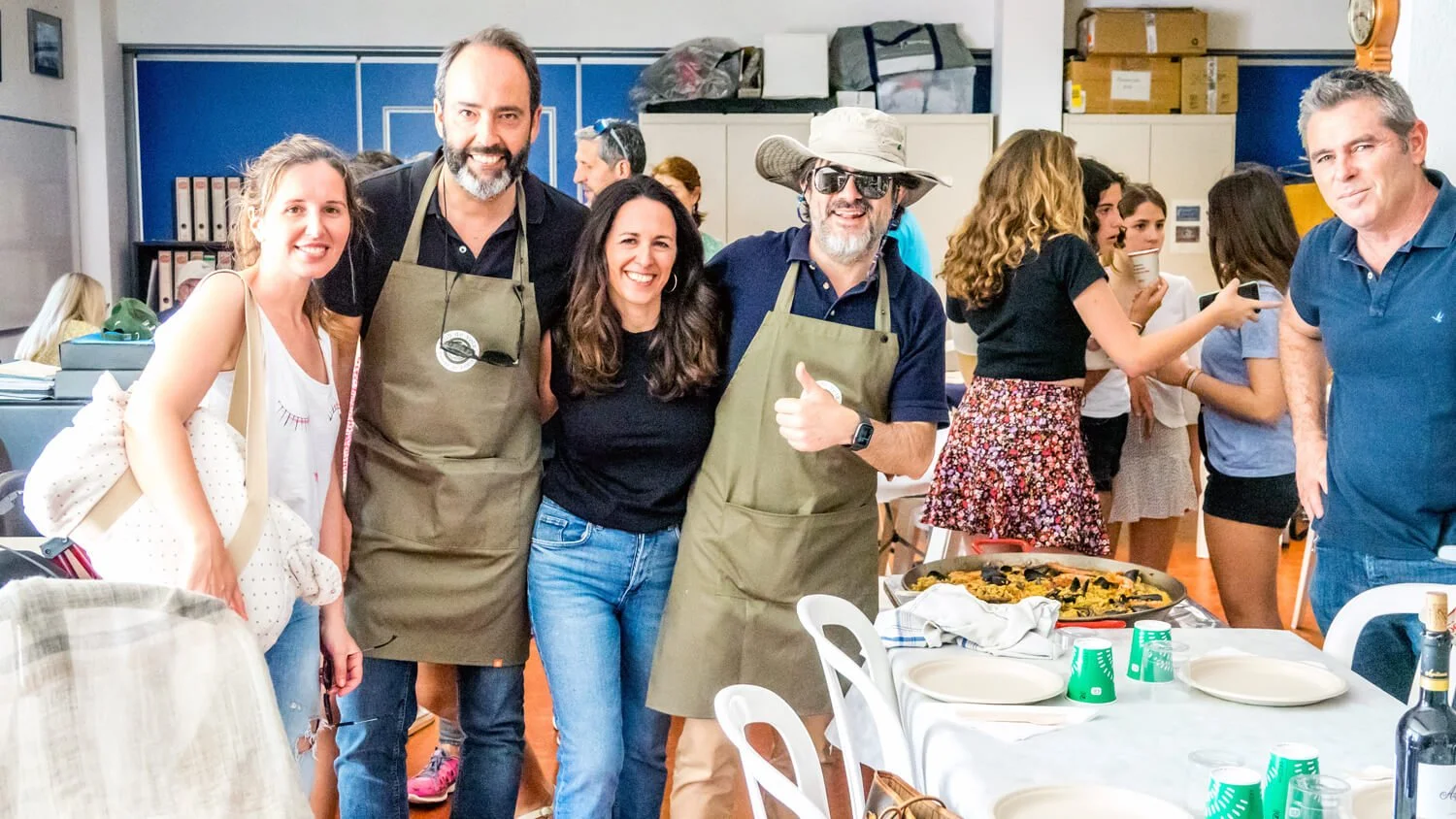 Grupo de personas en una celebración, algunos usando delantales, en una sala con mesas y comida, sonriendo y posando para la cámara.