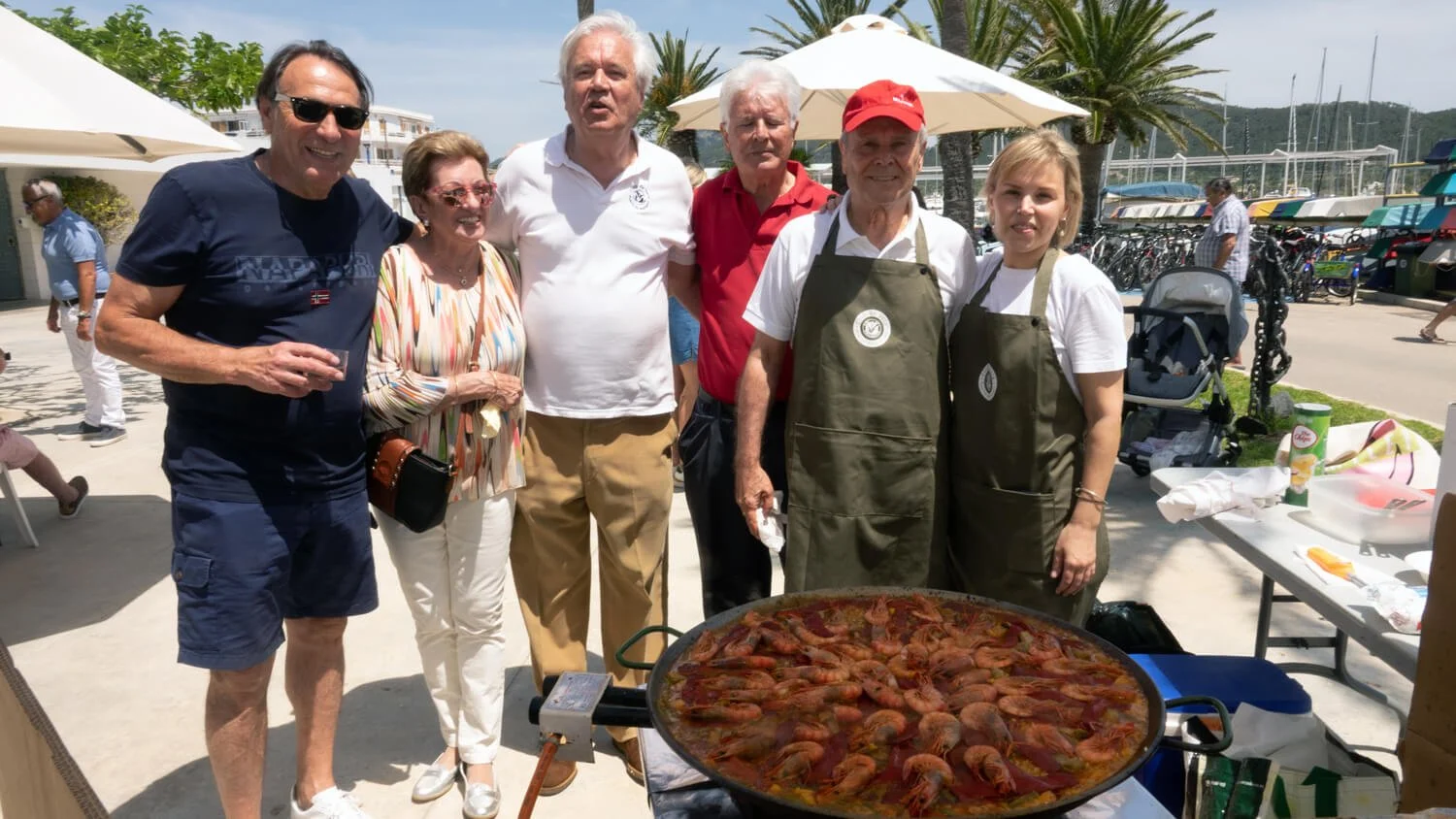 Un grupo de siete personas, algunos con delantales, posando junto a una paella con mariscos en una celebración al aire libre cerca del mar, con barcos y palmeras de fondo.