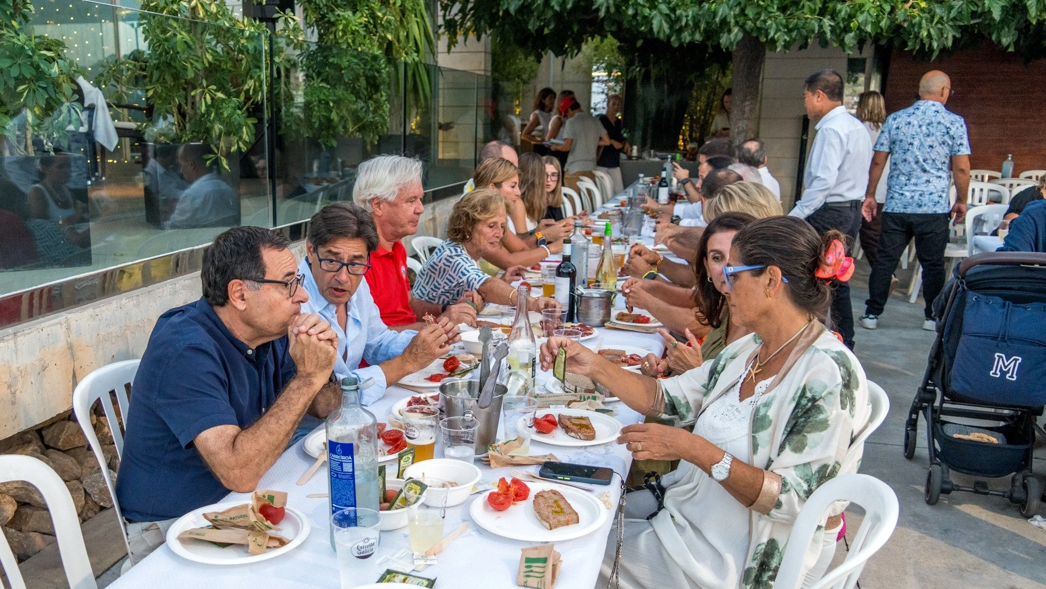 Grupo de personas de diferentes edades sentadas en una mesa al aire libre, disfrutando de una comida en un evento social.
