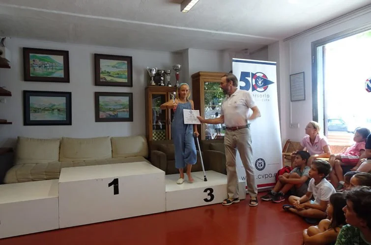 Persona en un podio recibe un diploma de otro hombre en un evento escolar, con niños sentados en el suelo y adultos en una sala decorada con cuadros y una vitrina con trofeos.