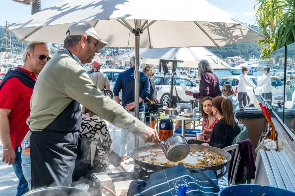 Hombre cocinando en una parrilla en un muelle con vista a yates, rodeado de personas en un ambiente al aire libre, con sombrillas y coches en el fondo.