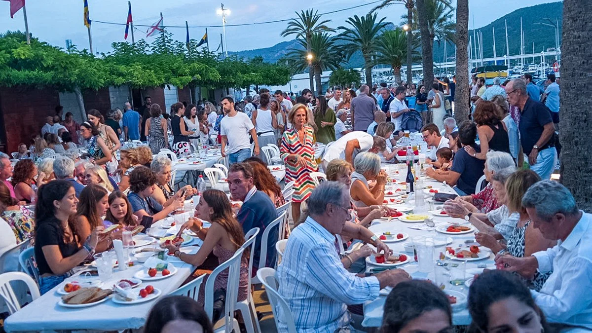 Imagen de una celebración al aire libre con muchas personas sentadas en mesas decoradas, disfrutando de comida y bebida, en un puerto con palmeras y montañas de fondo.