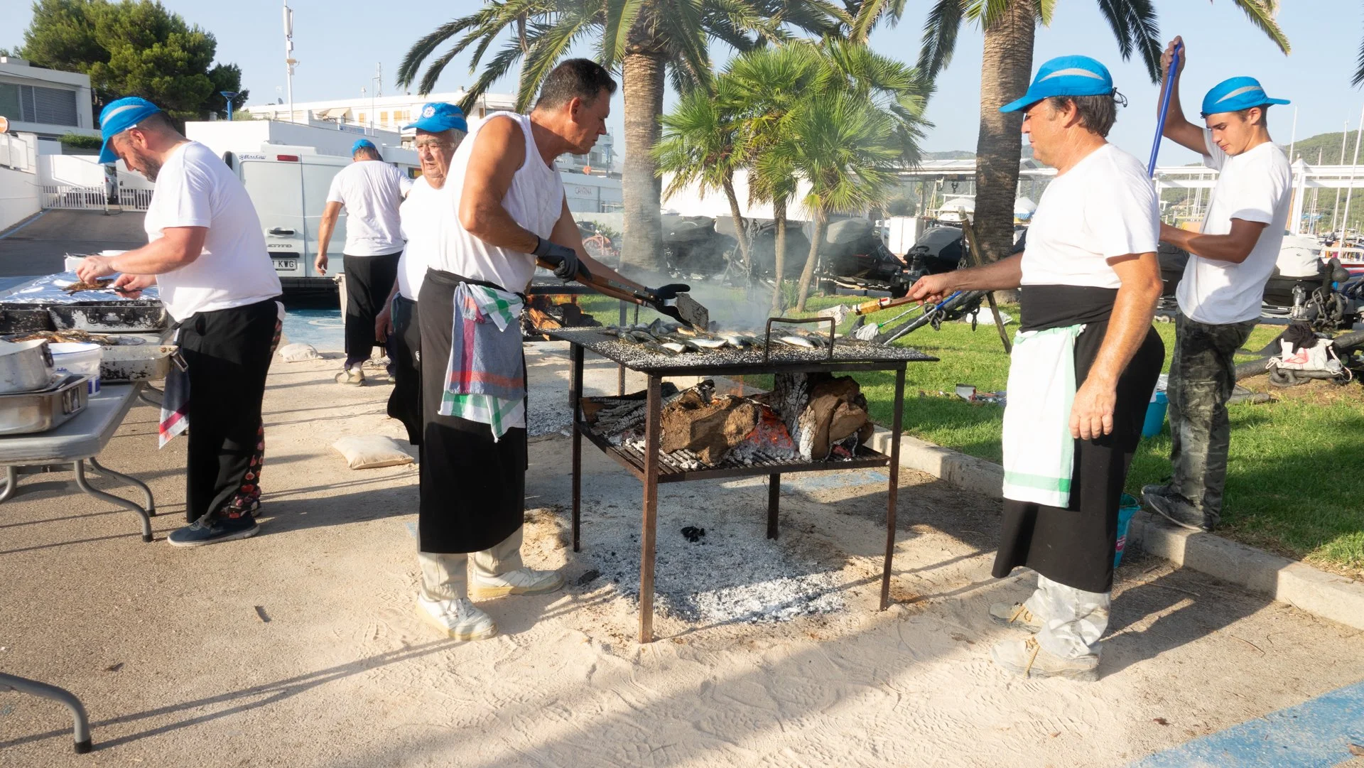 Grupo de personas cocinando pescado a la parrilla al aire libre junto a palmeras y junto a un área de parqueo en un día soleado.