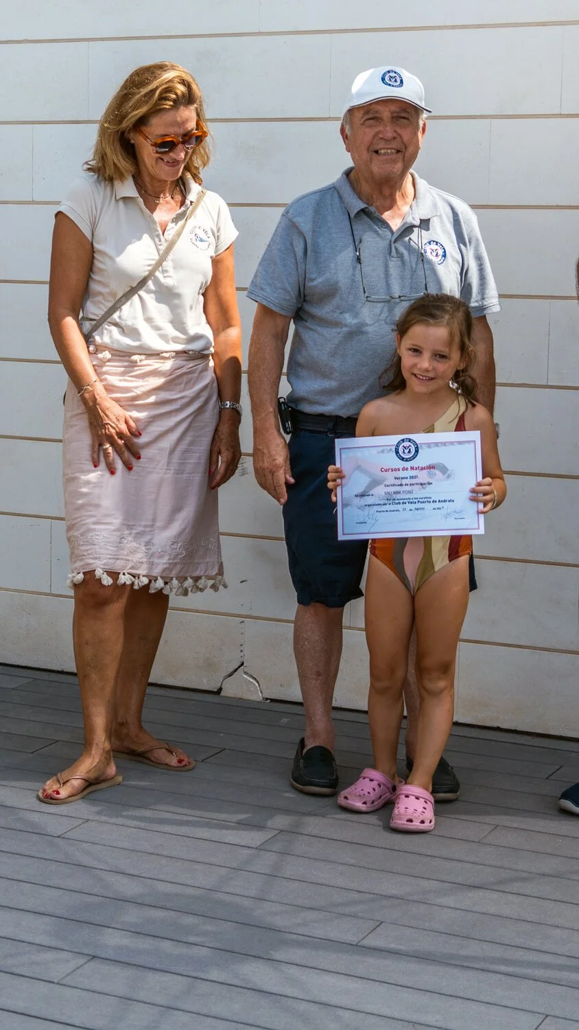 Niña en traje de baño recibe un certificado de participación en un curso de natación, acompañada por un hombre y una mujer en un evento al aire libre.