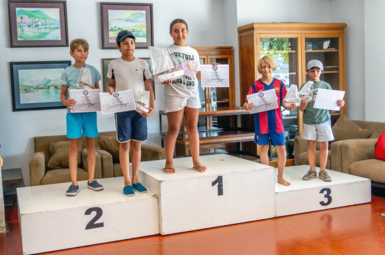 Niños en un podio de premios en un evento, con certificados y una pequeña embarcación de vela, dentro de una sala con cuadros y muebles de madera.