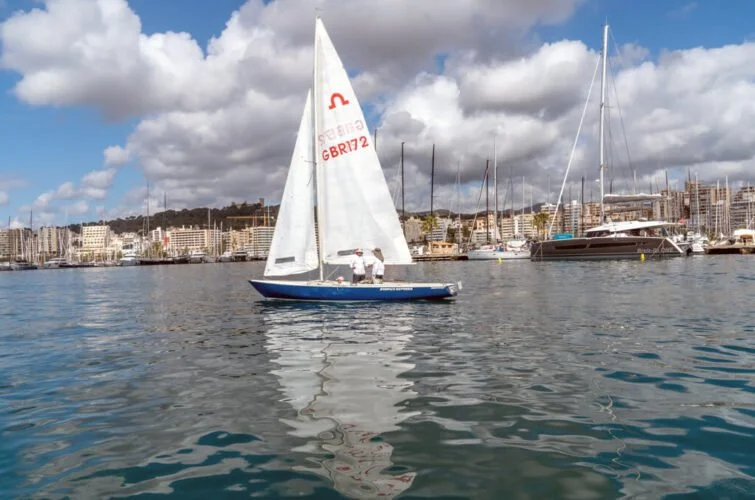 Un velero navegando en un puerto con edificios y barcos en el fondo, bajo un cielo nublado.