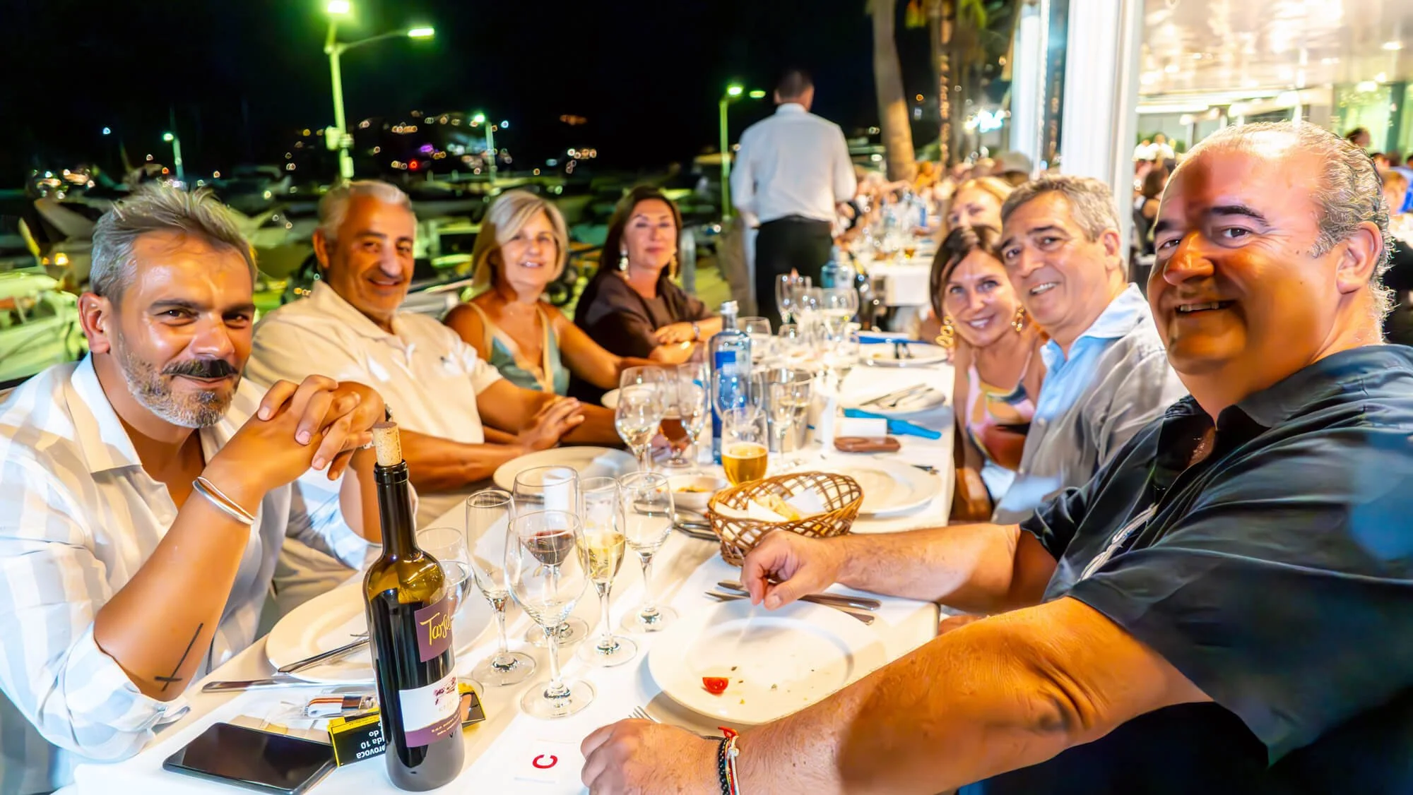 Grupo de personas sentadas en una mesa en un restaurante al aire libre durante la noche, disfrutando de comida y bebidas, con un fondo de yates y luces en un puerto.