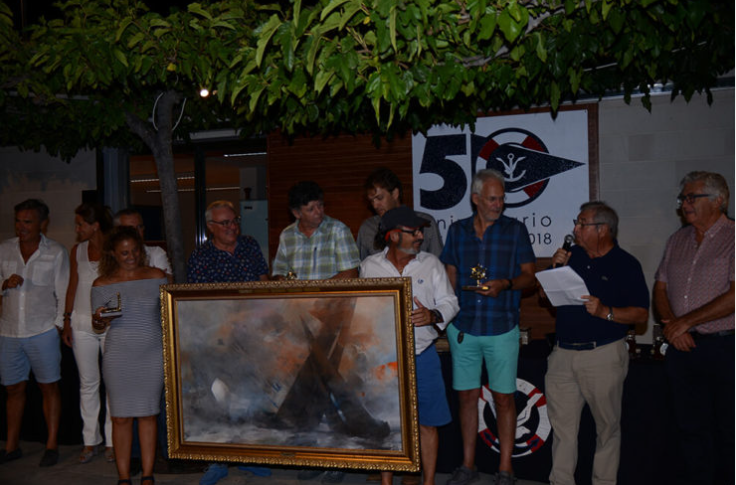 Grupo de personas en una ceremonia, con un hombre que lee un discurso y una mujer que sostiene un cuadro de estilo abstracto, en un evento con un cartel de 50 años en el fondo.