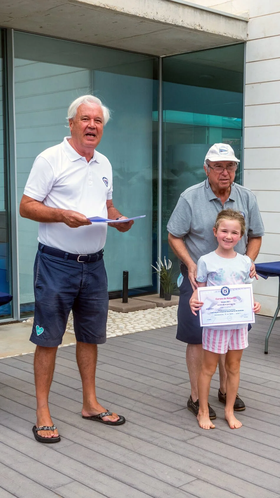 Una niña recibe un certificado de participación en una ceremonia al aire libre, acompañada por un hombre mayor, mientras otro hombre con cabello canoso y con camisa y pantalones cortos está a su lado, leyendo un documento.