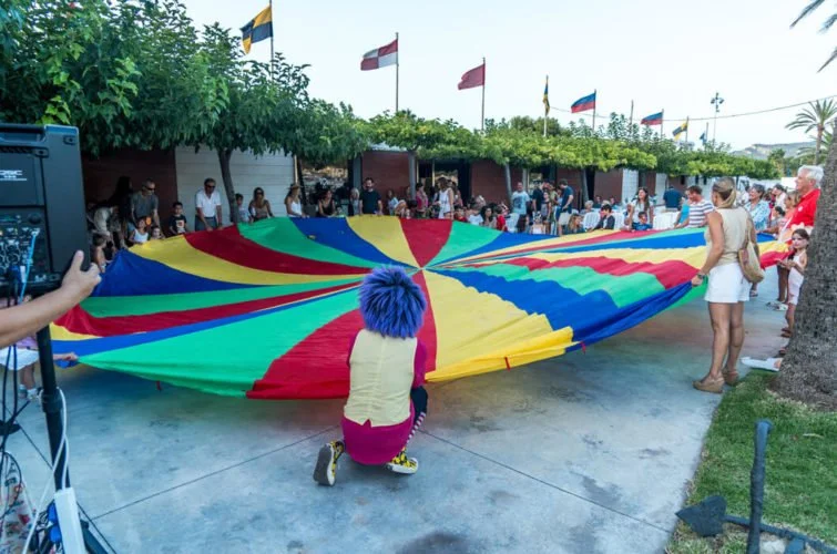 Grupo de personas participando en un evento con un paracaídas de colores en un espacio al aire libre, con banderas en el fondo.