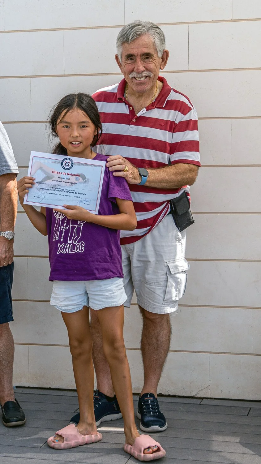 Una niña joven recibe un certificado de natación en una ceremonia al aire libre, mientras un hombre mayor, probable instructor o familiar, la felicita y la apoya.