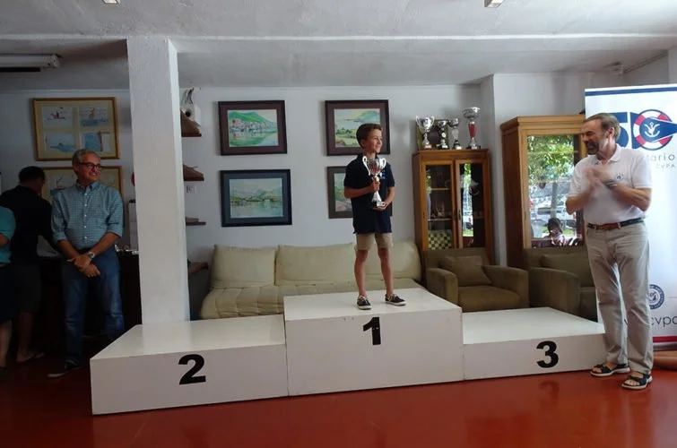 Niño en primer lugar en una ceremonia de premiación, con un trofeo, en un podio de tres niveles, en un salón con decoraciones y personas observando.