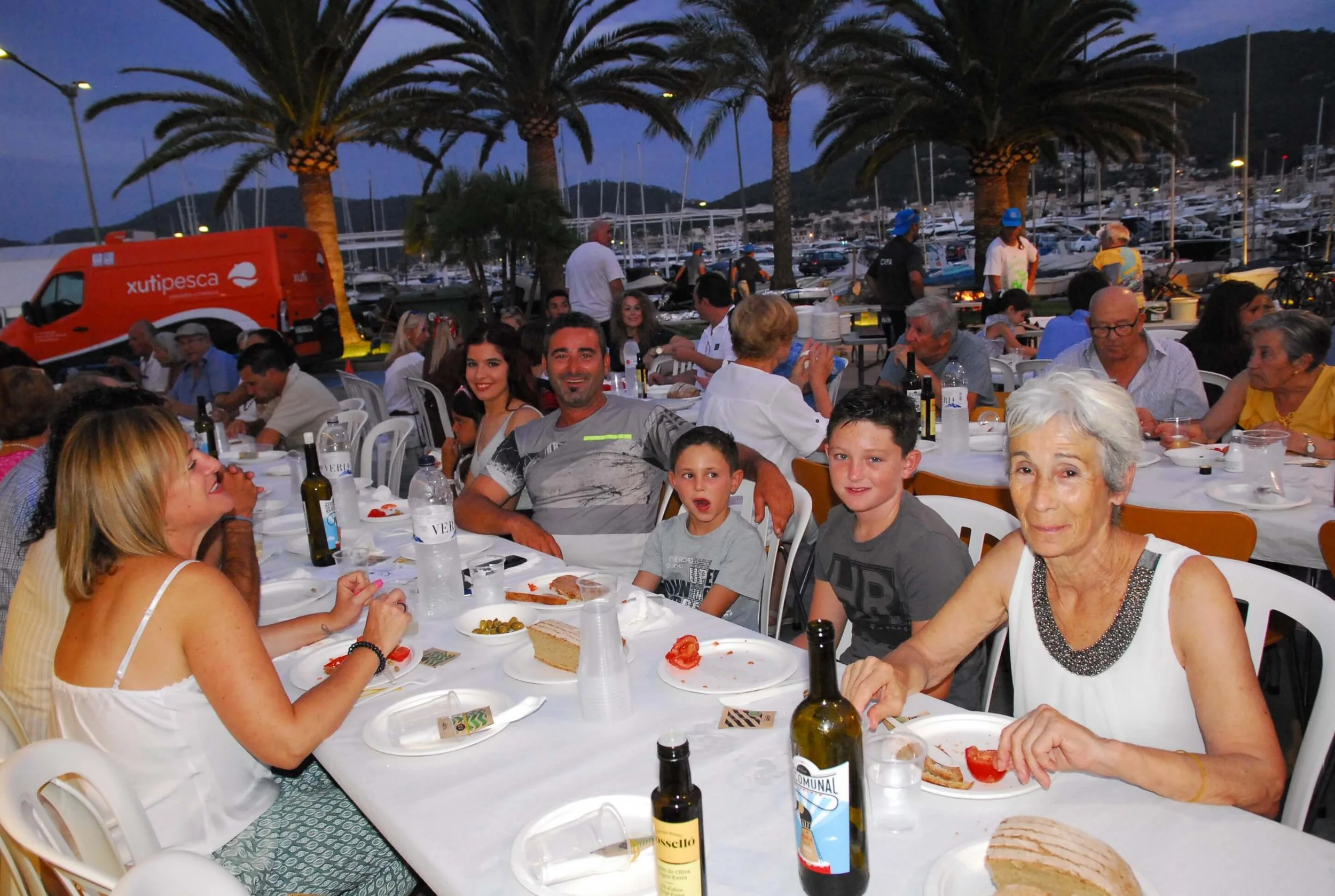 Imagen de una reunión familiar o social en una terraza junto al puerto con montañas al fondo, durante la noche. Hay personas sentadas en mesas disfrutando comida y bebida, algunos niños y adultos felices. En el fondo hay palmeras, muchos yates y barc
