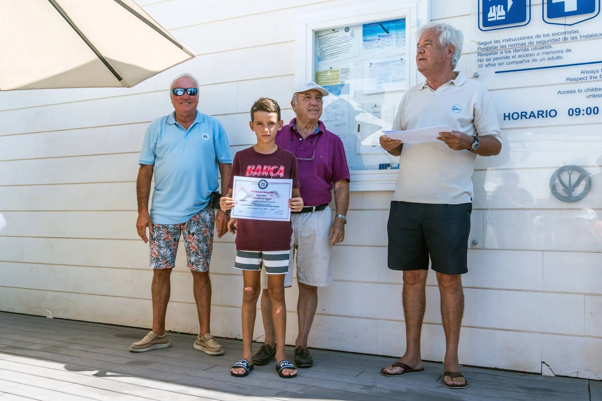Un grupo de cinco personas, cuatro adultos y un niño, de piel clara, están de pie frente a un instituto de natación, con un cartel azul y un aviso en la pared, en un día soleado. El niño sostiene un certificado de natación; los adultos parecen ser en