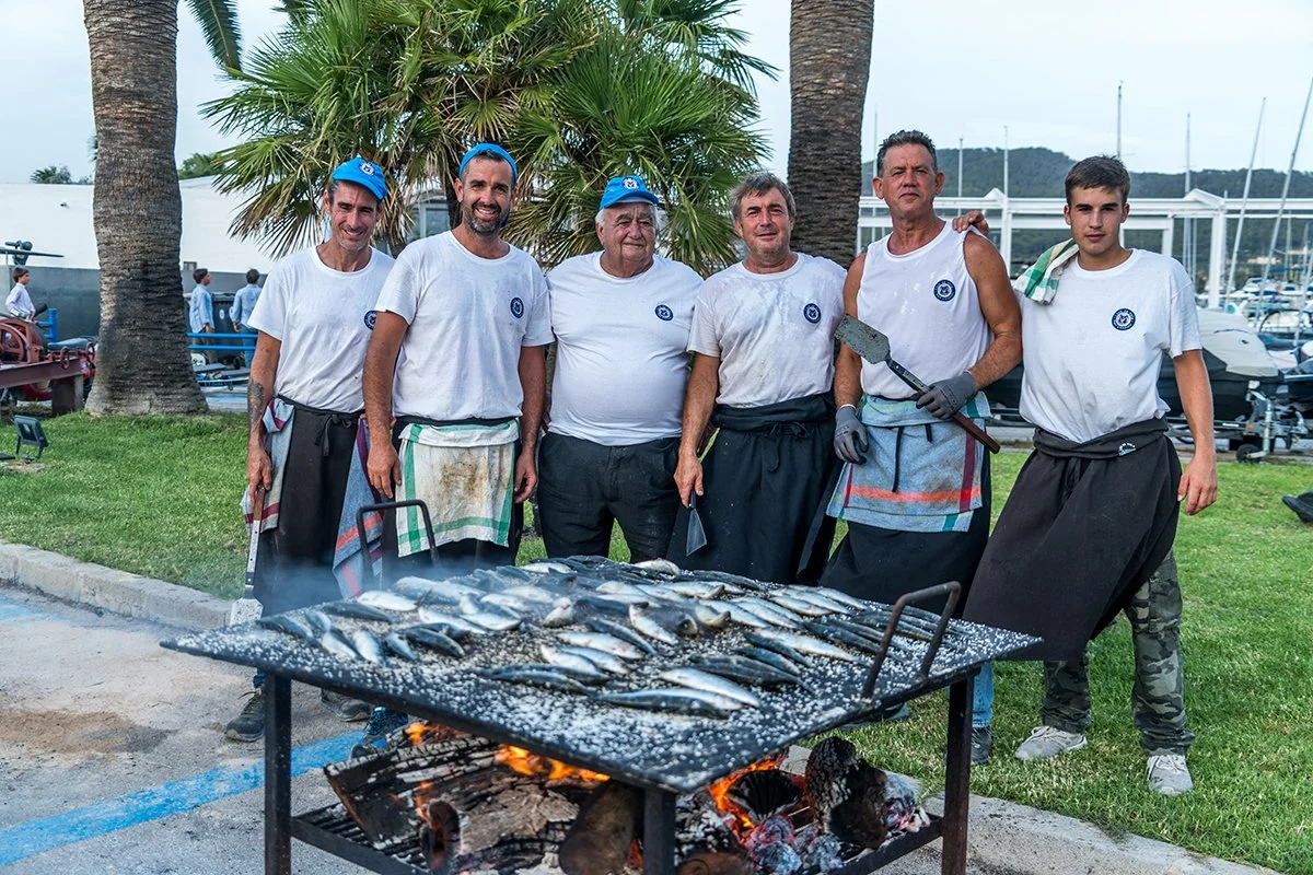 Grupo de seis personas posando detrás de una parrilla con pescado asado, en un parque con palmeras y yates en el fondo.