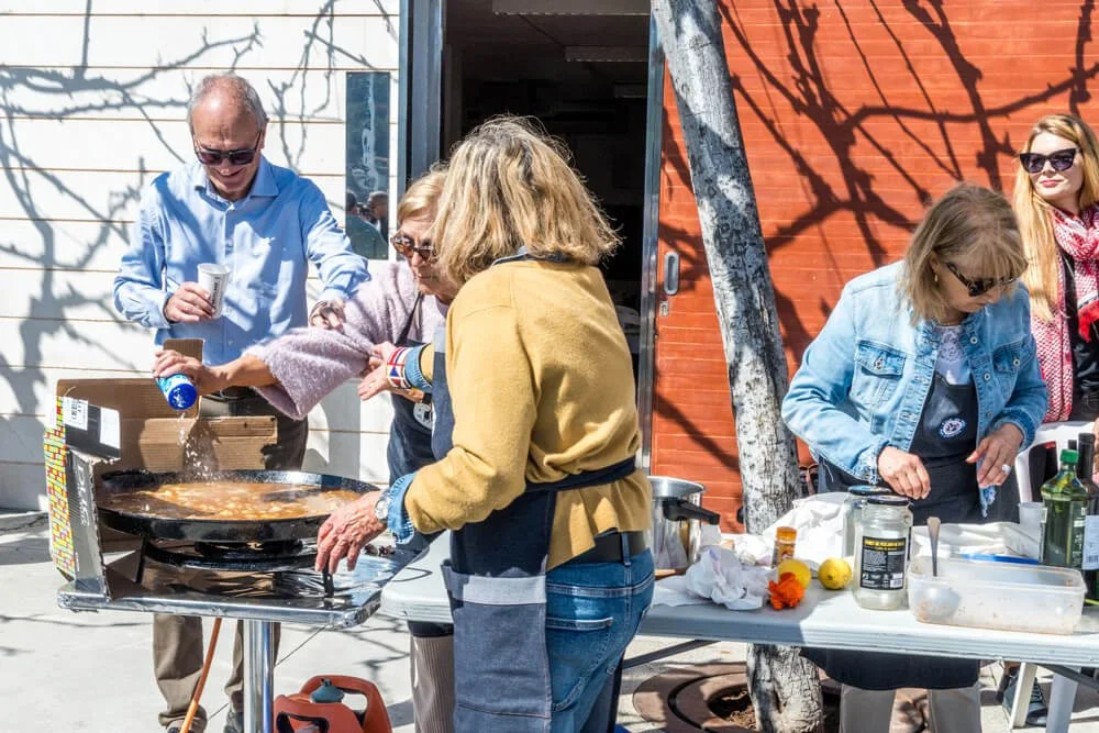 Varias personas cocinando y sirviendo comida en un área al aire libre con paredes de madera y sombra de árbol.