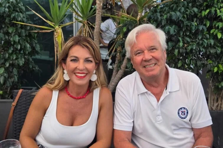 Una mujer y un hombre sonrientes sentados en una mesa al aire libre, rodeados de plantas y decoración tropical.