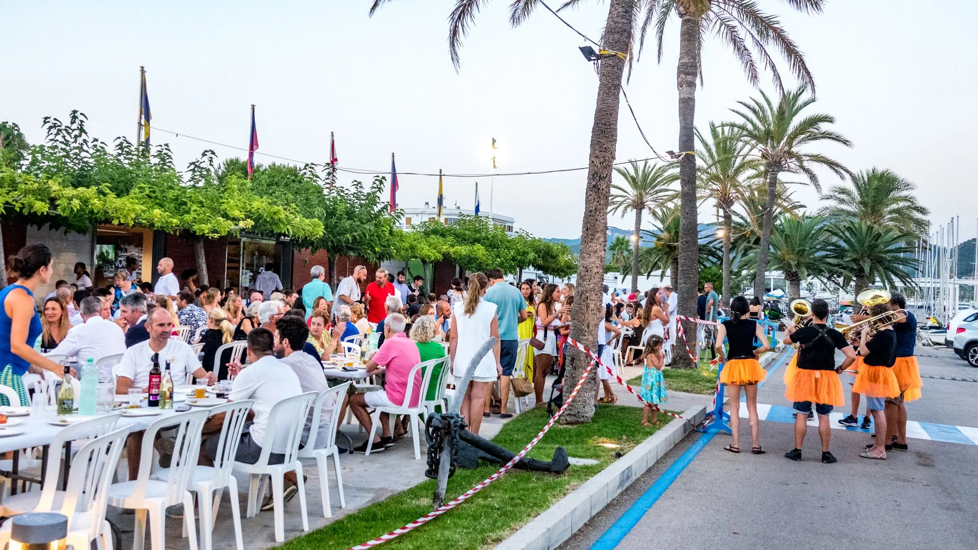Una reunión o celebración al aire libre con muchas personas sentadas en mesas y otras de pie, y un grupo de músicos con instrumentos musicales vistiendo en disfraces de color naranja. Hay palmeras y banderas en el fondo.