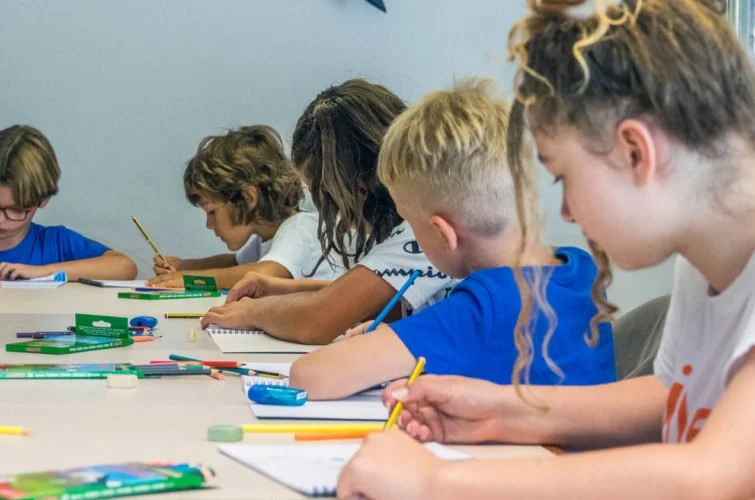 Grupo de niños y una niña sentados en una mesa, escribiendo y dibujando en papeles con lápices y útiles escolares.