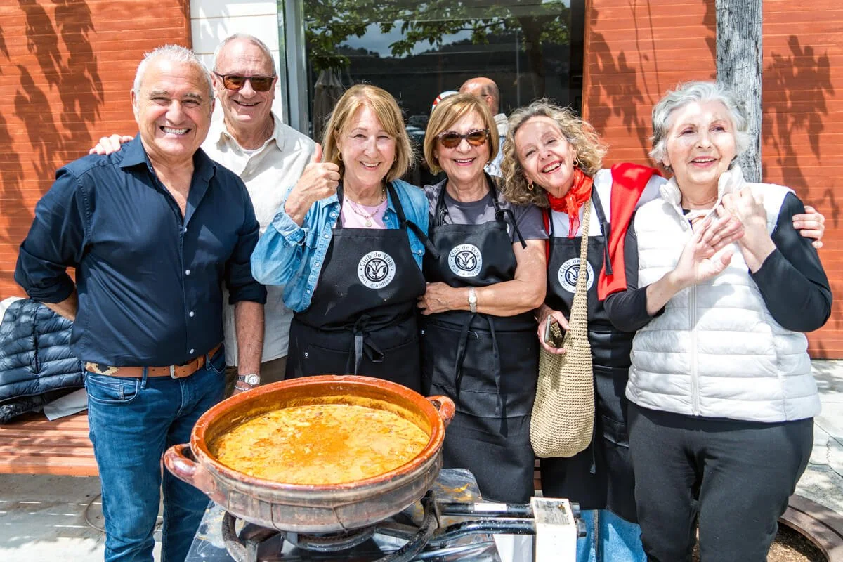Grupo de personas mayores riendo y sonriendo junto a una olla de comida en una reunión al aire libre, con fondo de muro de ladrillo y árboles