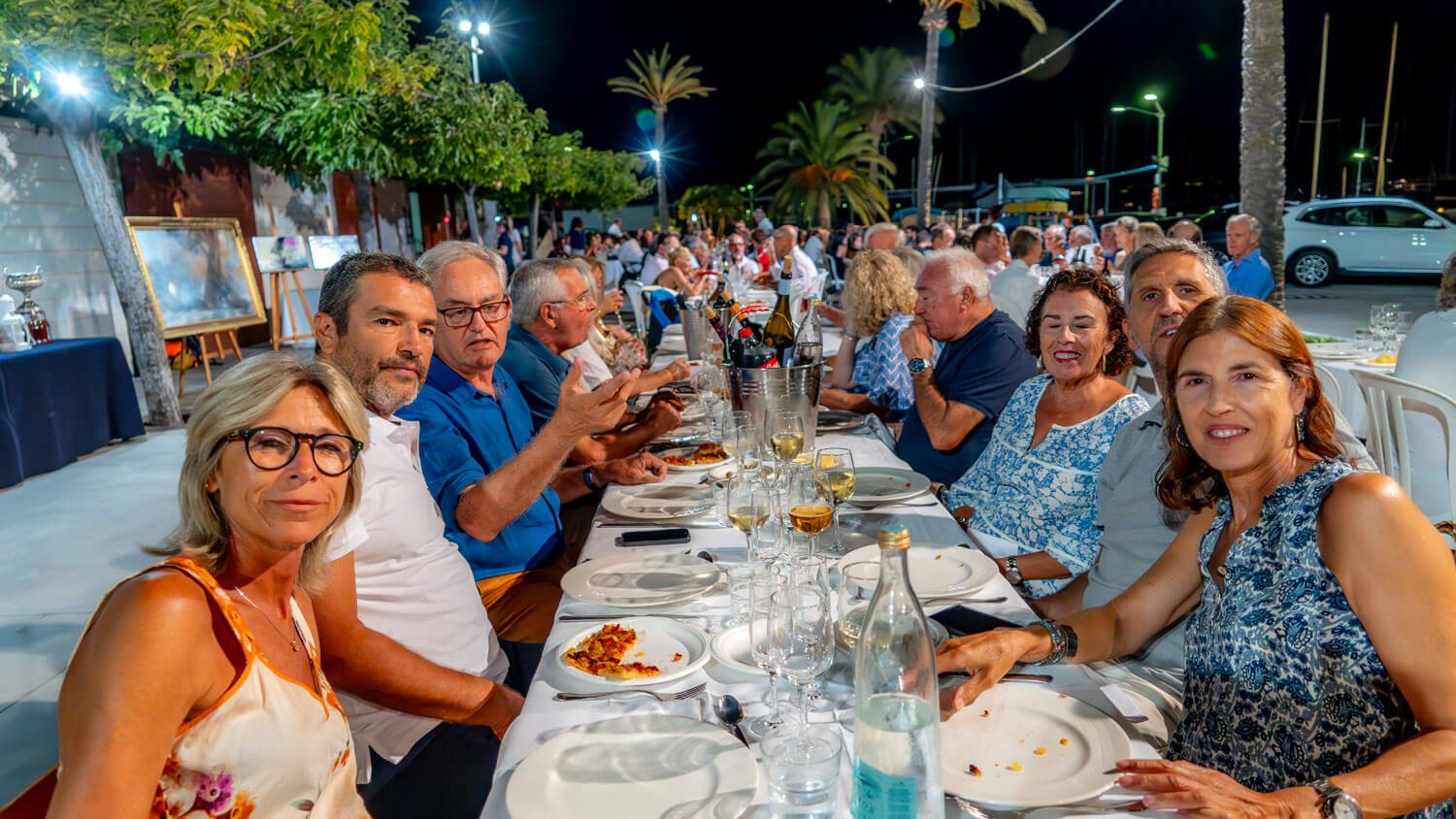 Grupo de personas sentadas en una mesa larga al aire libre de noche, comiendo y socializando en un evento festivo con decoración y árboles en el fondo.