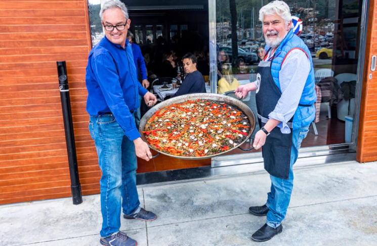 Dos hombres sosteniendo una pizza gigante frente a un restaurante, uno con gafas y camisa azul, el otro con barba y chaqueta azul. Interior del restaurante visible a través de la puerta de vidrio.