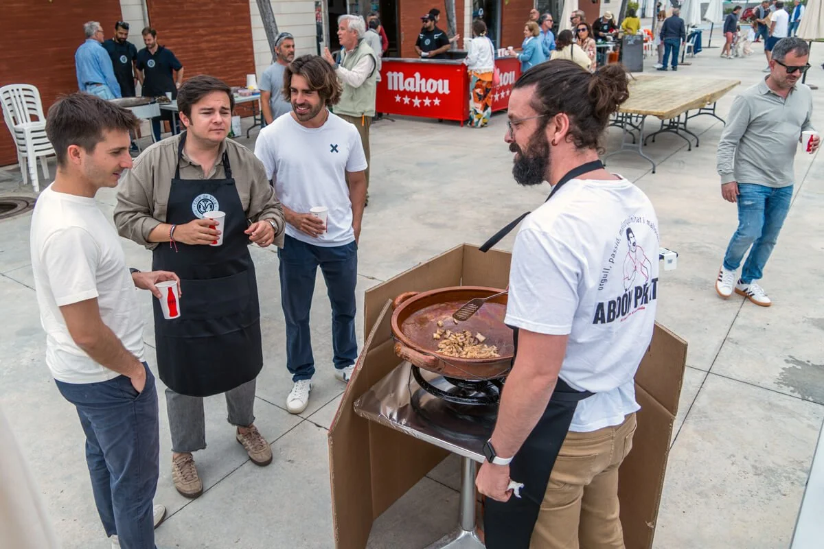 Hombres disfrutando en una feria o evento al aire libre, uno de ellos sirviendo un plato de comida caliente en un recipiente grande, mientras otros observan y conversan, con varias mesas y puestos en el fondo.