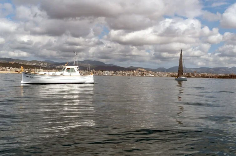 Barco y velero en un cuerpo de agua con ciudad y montañas al fondo, cielo nublado.