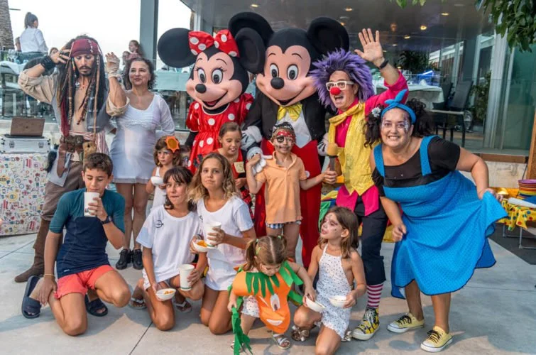 Grupo de niños y adultos con disfraces, acompañados por Mickey y Minnie Mouse y personajes de payasos, en una celebración al aire libre.