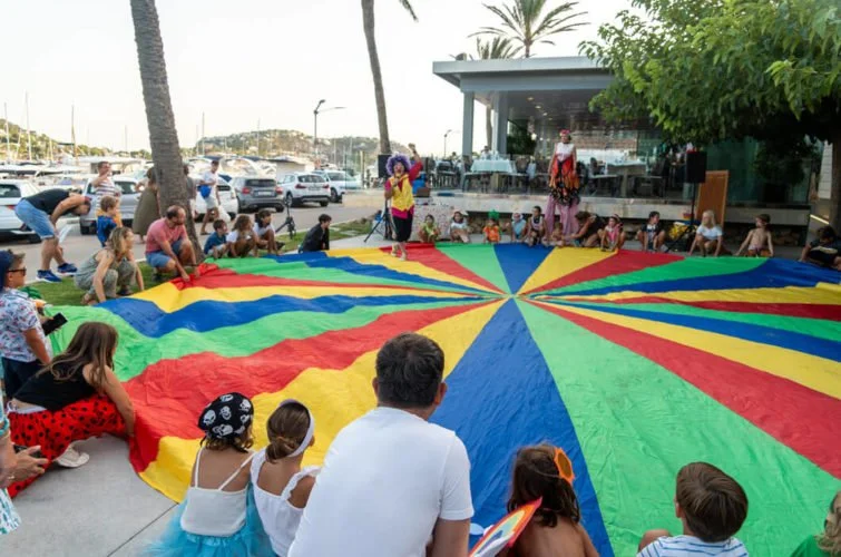 Niños y adultos participando en una actividad con un paracaídas colorido, en un espacio al aire libre cerca del mar, con palmeras y un escenario con payasos y personas disfrazadas.