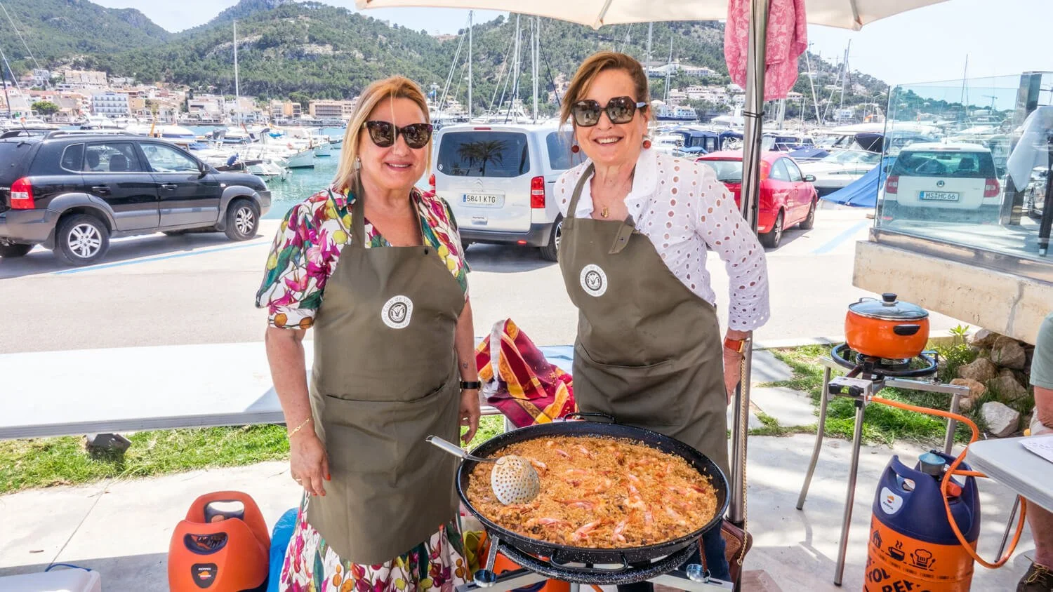 Dos mujeres cocinando comida en una parque en un puerto con botes y autos en el fondo.