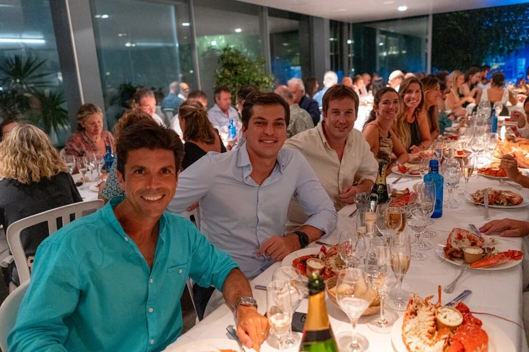 Grupo de personas en una cena formal, sentados a una mesa larga con comida y bebidas, en un ambiente elegante y bien iluminado.