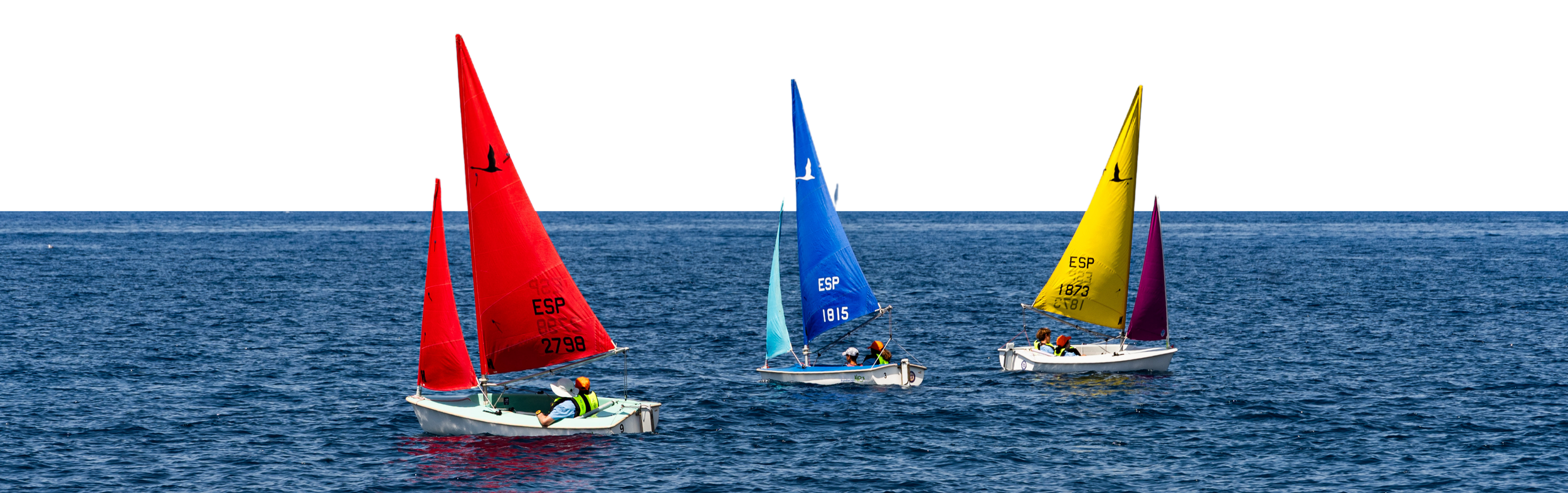 Tres pequeños barcos de vela con velas de colores rojo, azul y amarillo navegando en el mar, con una estructura en el fondo y varios niños en los barcos.
