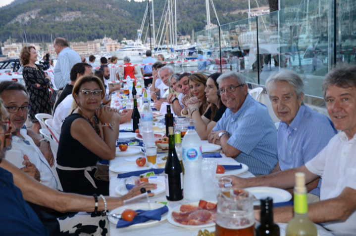 Grupo de personas sentadas a una mesa al aire libre en un evento social, con barcos y montañas de fondo.