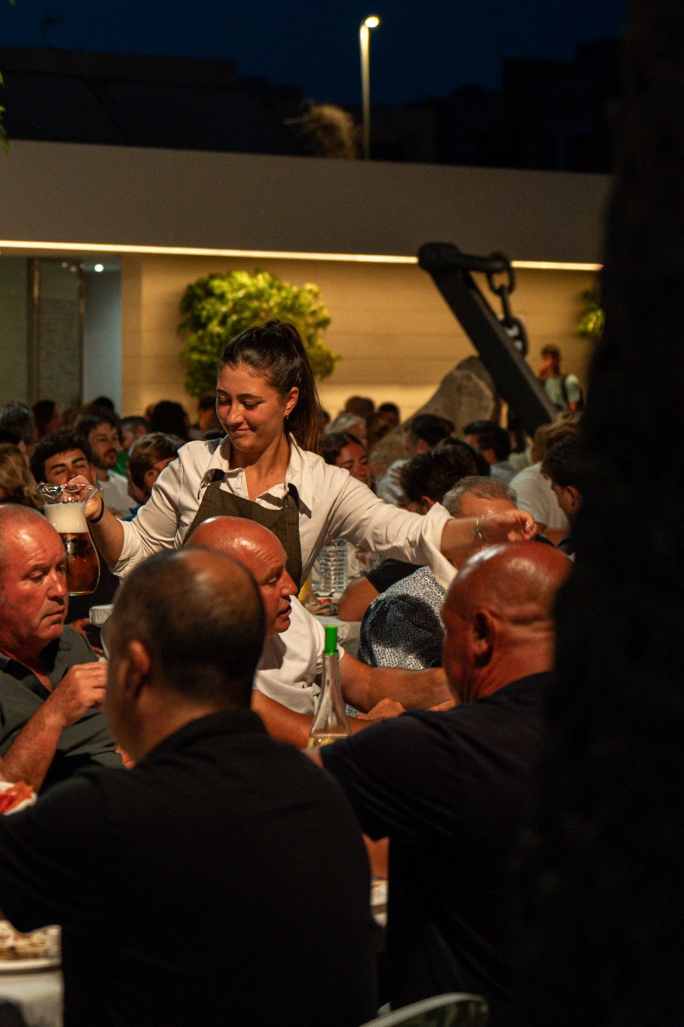 Una mujer sirviendo comida o bebida en una reunión nocturna con muchas personas sentadas en mesas. Parece un evento social o festival, en un entorno al aire libre con iluminación artificial.
